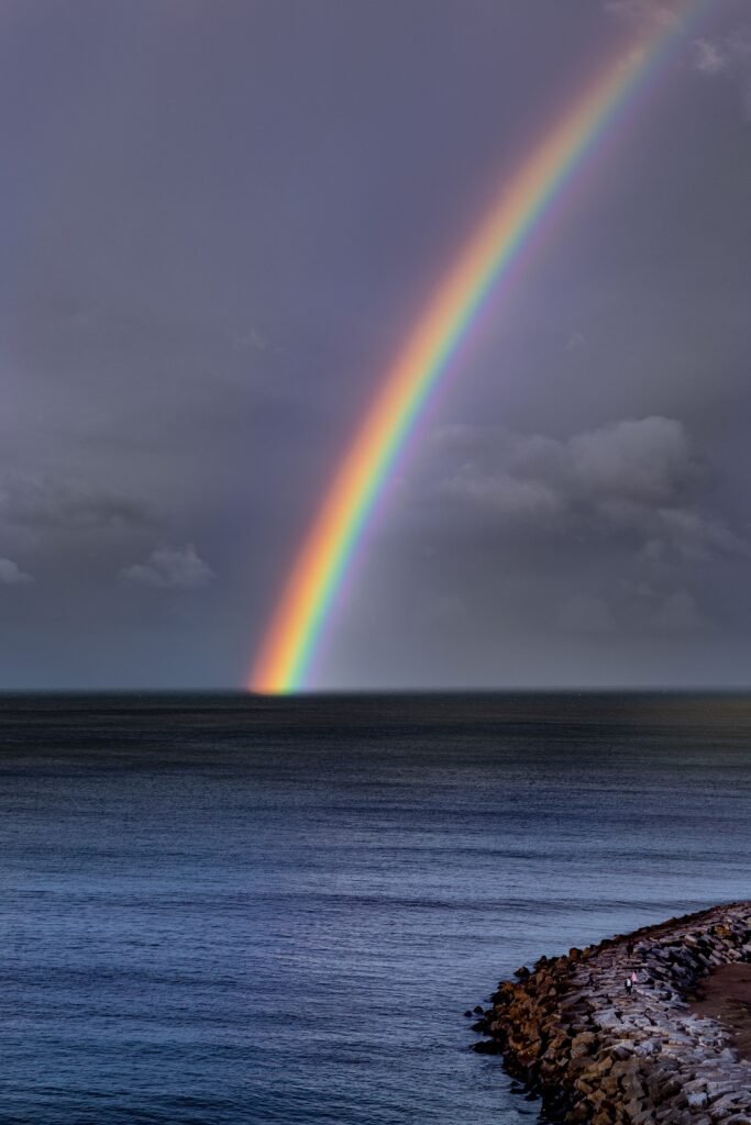 Arcoíris que nace sobre el mar bajo un cielo gris de tormenta, junto a un espigón de rocas