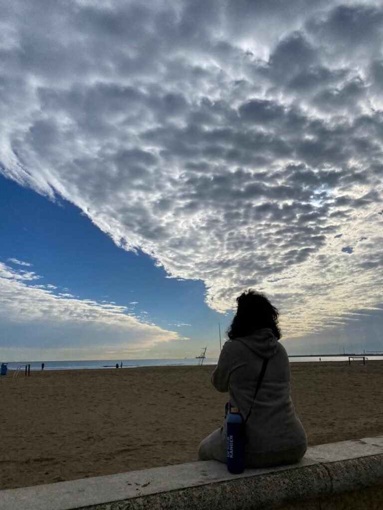 Ana Belén Úbeda García sentada de espaldas mirando al mar bajo un cielo lleno de nubes