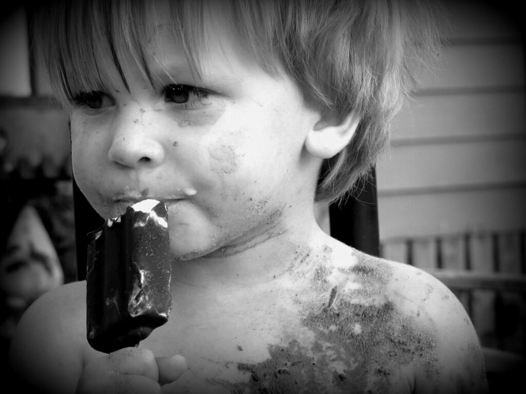 Niño pequeño comiendo un helado de chocolate, con la cara y los hombros manchados de arena, en una foto en blanco y negro